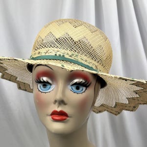 May include: A straw hat with a wide, scalloped brim. The hat features a woven design on the crown and a green ribbon detail. The brim has a decorative edge, and the hat is displayed on a mannequin head.