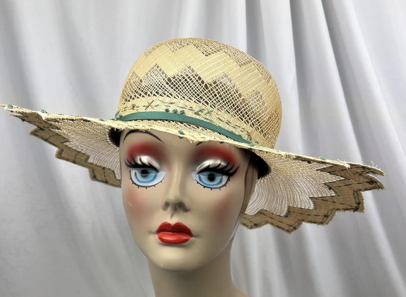 May include: A straw hat with a wide, scalloped brim. The hat features a woven design on the crown and a green ribbon detail. The brim has a decorative edge, and the hat is displayed on a mannequin head.