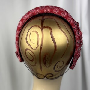 May include: A red headband with a repeating floral pattern and small embellishments. The headband is displayed on a gold-painted mannequin head.