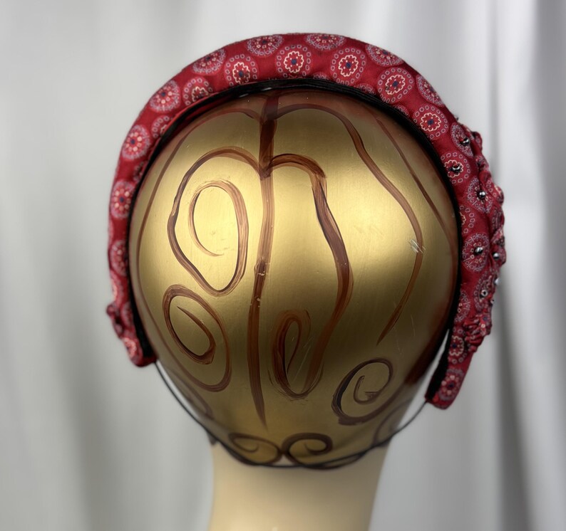 May include: A red headband with a repeating floral pattern and small embellishments. The headband is displayed on a gold-painted mannequin head.