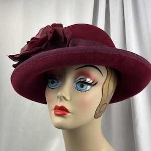 May include: A deep burgundy felt hat featuring a wide brim and a matching ribbon band. A large, layered fabric flower in a coordinating dark red shade is attached to the side of the crown, adding texture to the smooth, structured felt material.