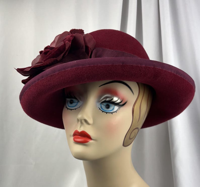 May include: A deep burgundy felt hat featuring a wide brim and a matching ribbon band. A large, layered fabric flower in a coordinating dark red shade is attached to the side of the crown, adding texture to the smooth, structured felt material.