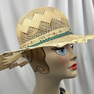 May include: A straw hat with a wide, scalloped brim and a decorative green band. The hat features a woven design on the crown and a delicate, open weave throughout. The hat is displayed on a mannequin head.