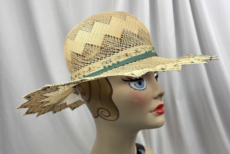May include: A straw hat with a wide, scalloped brim and a decorative green band. The hat features a woven design on the crown and a delicate, open weave throughout. The hat is displayed on a mannequin head.