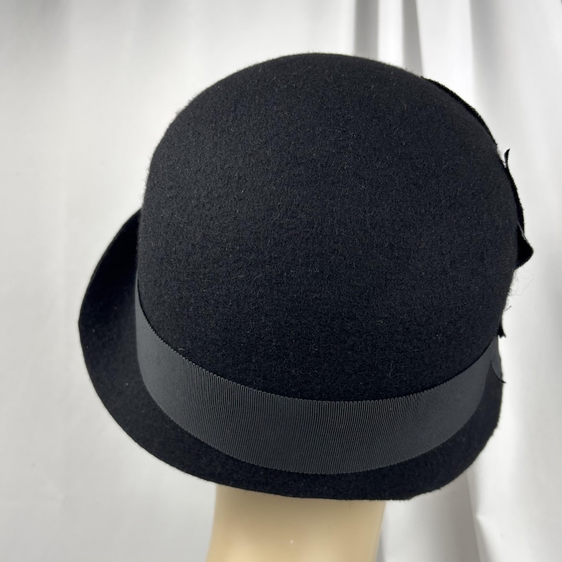 May include: A solid black felt cloche hat featuring a rounded crown and a narrow, slightly upturned brim. A matching black grosgrain ribbon band encircles the base of the crown, and a decorative black fabric flower is attached to the side.