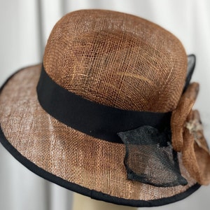 Pu&ograve; includere: Un cappello di paglia marrone con un nastro nero e un fiocco di fiori marroni e neri.