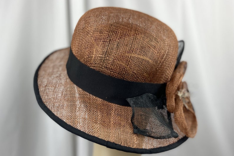 Pu&ograve; includere: Un cappello di paglia marrone con un nastro nero e un fiocco di fiori marroni e neri.