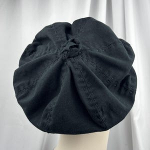 May include: A black beret featuring a central knot detail at the crown and radiating rows of decorative tonal running stitches. The fabric is gathered into soft folds, creating a rounded silhouette that sits atop a smooth, neutral-toned base.