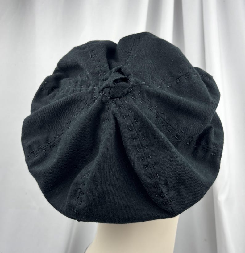 May include: A black beret featuring a central knot detail at the crown and radiating rows of decorative tonal running stitches. The fabric is gathered into soft folds, creating a rounded silhouette that sits atop a smooth, neutral-toned base.