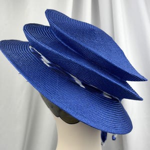 K&ouml;nnte beinhalten: Ein leuchtend k&ouml;nigsblauer Strohhut mit einem mehrstufigen, geschichteten Design, einer runden Krone und einer breiten Krempe. Ein d&uuml;nnes, durchscheinendes Band mit wei&szlig;-blauem Muster ist um die Basis der Krone gewickelt und setzt einen Kontrast.