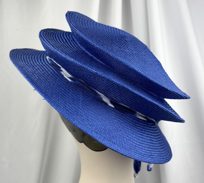 K&ouml;nnte beinhalten: Ein leuchtend k&ouml;nigsblauer Strohhut mit einem mehrstufigen, geschichteten Design, einer runden Krone und einer breiten Krempe. Ein d&uuml;nnes, durchscheinendes Band mit wei&szlig;-blauem Muster ist um die Basis der Krone gewickelt und setzt einen Kontrast.
