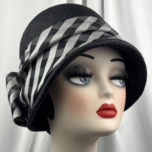 May include: Black cloche hat with a wide brim and a black and white checkered ribbon. The hat is made of woven material and has a bow on the side. A stylish accessory for any occasion.