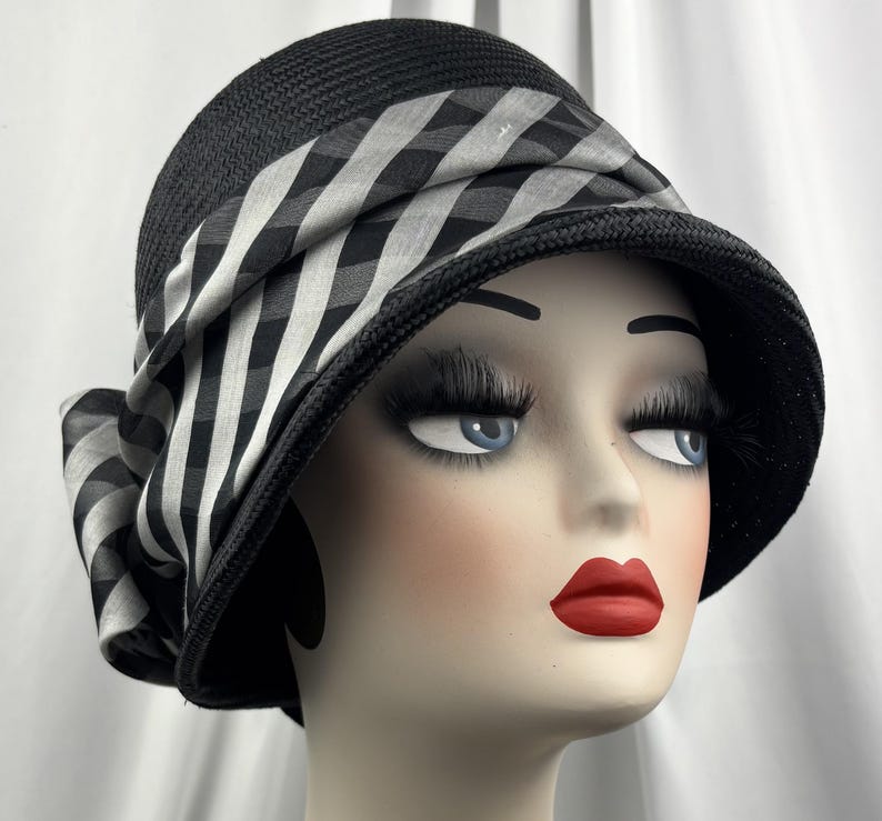 May include: Black cloche hat with a wide brim and a black and white checkered ribbon. The hat is made of woven material and has a bow on the side. A stylish accessory for any occasion.
