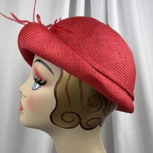 May include: A bright red woven straw hat with a rounded crown and a slightly upturned brim. The hat is decorated with a cluster of thin, delicate red feathers attached to the side, adding a textured detail to the solid red woven material.