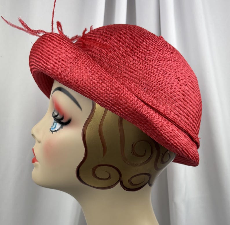 May include: A bright red woven straw hat with a rounded crown and a slightly upturned brim. The hat is decorated with a cluster of thin, delicate red feathers attached to the side, adding a textured detail to the solid red woven material.