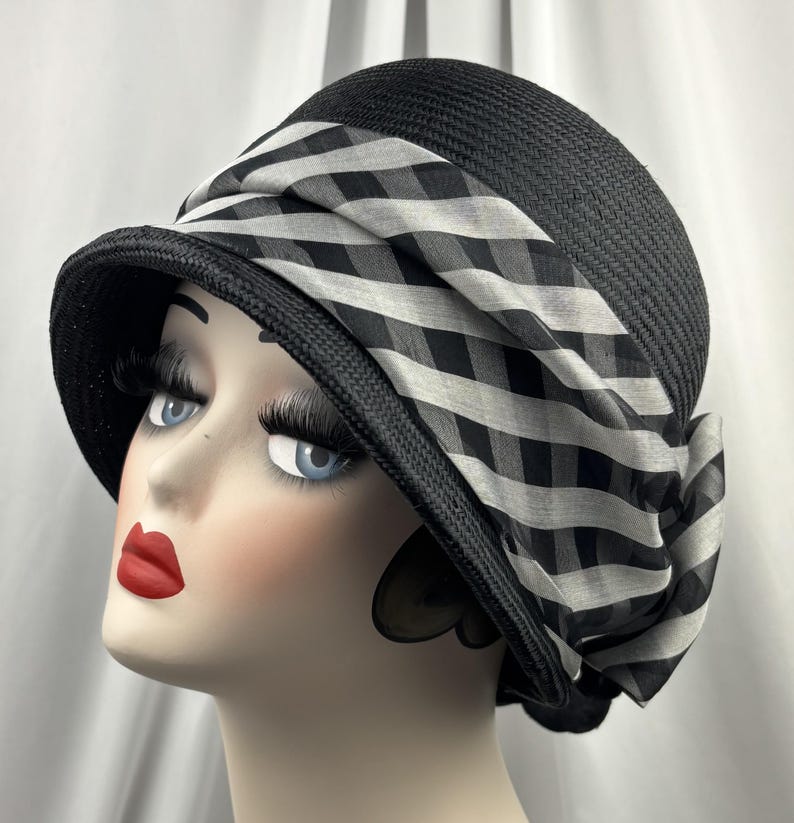 May include: Black cloche hat with a wide brim and a black and white checked ribbon. The hat is made of woven material and has a rounded crown. The ribbon is draped around the hat and tied in a bow.
