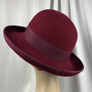 May include: A deep burgundy felt hat featuring a rounded crown and a wide, slightly upturned brim. The hat is accented with a matching dark red grosgrain ribbon band around the base of the crown and a coordinating ribbon trim along the brim edge.