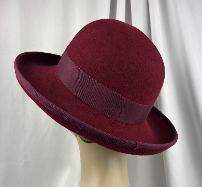 May include: A deep burgundy felt hat featuring a rounded crown and a wide, slightly upturned brim. The hat is accented with a matching dark red grosgrain ribbon band around the base of the crown and a coordinating ribbon trim along the brim edge.