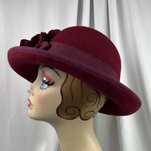 May include: A deep burgundy felt hat featuring a wide brim and a matching ribbon band around the crown. A large, layered fabric flower in the same dark red tone is attached to the side of the hat, resting above the brim and ribbon detail.