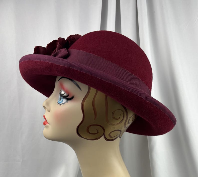 May include: A deep burgundy felt hat featuring a wide brim and a matching ribbon band around the crown. A large, layered fabric flower in the same dark red tone is attached to the side of the hat, resting above the brim and ribbon detail.