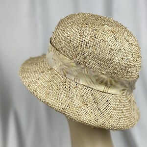 Puede incluir: Un sombrero de paja de ala ancha con una cinta estampada. El sombrero es de color beige claro y tiene una textura tejida.