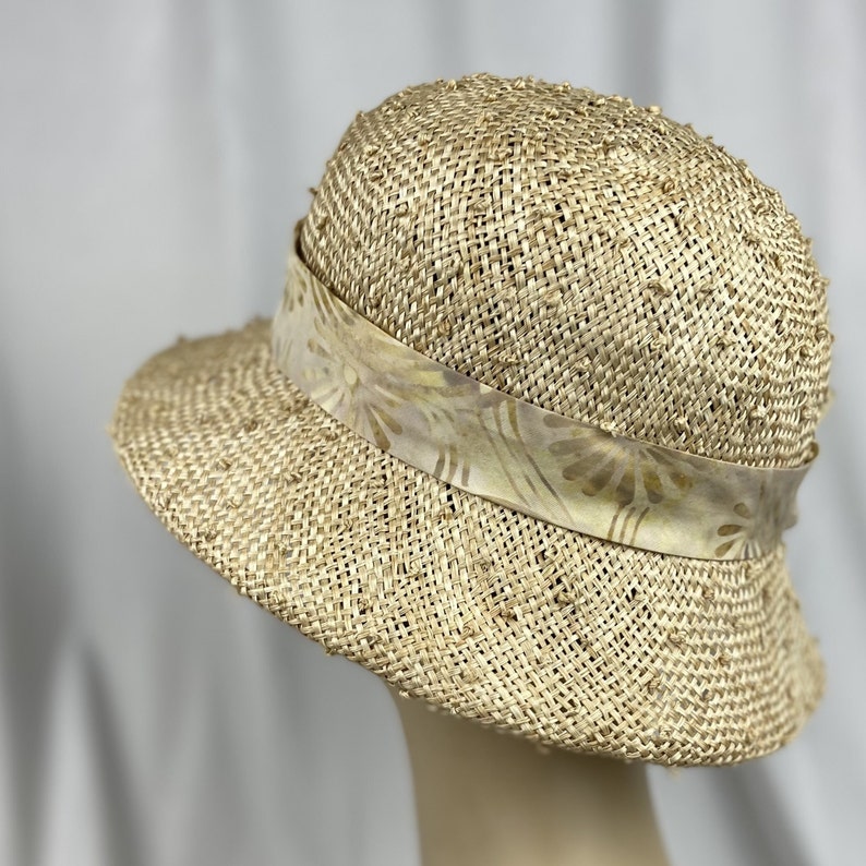 Puede incluir: Un sombrero de paja de ala ancha con una cinta estampada. El sombrero es de color beige claro y tiene una textura tejida.