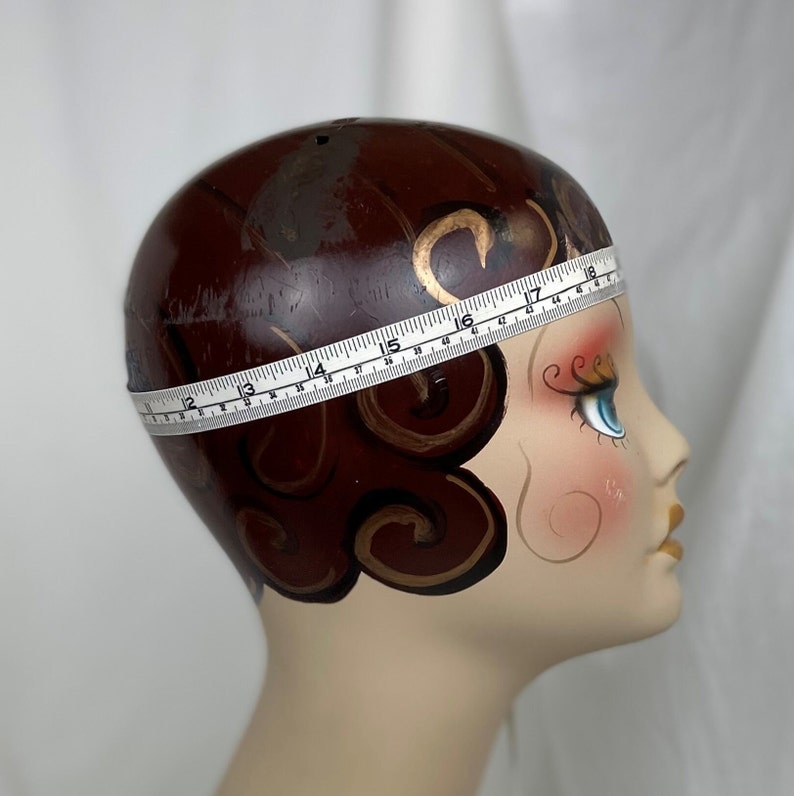 May include: A white measuring tape is wrapped around a mannequin head with brown hair and gold painted details. The tape reads 17 inches.