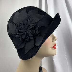 May include: A black felt cloche hat featuring a large, layered floral embellishment made from black grosgrain ribbon. The hat has a rounded crown and a small, asymmetrical brim that curves upward on one side, creating a structured silhouette.