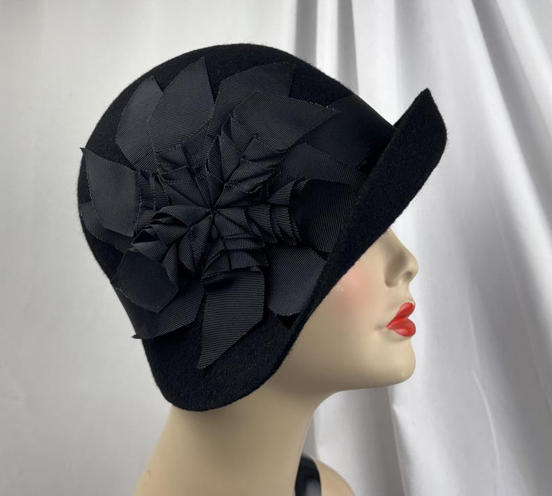 May include: A black felt cloche hat featuring a large, layered floral embellishment made from black grosgrain ribbon. The hat has a rounded crown and a small, asymmetrical brim that curves upward on one side, creating a structured silhouette.