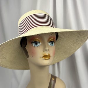 Pu&ograve; includere: Un cappello di paglia a tesa larga con corona e tesa color crema. Il cappello presenta una fascia a strisce bordeaux e bianche alternate intorno alla corona. Il cappello &egrave; esposto su una testa di manichino.