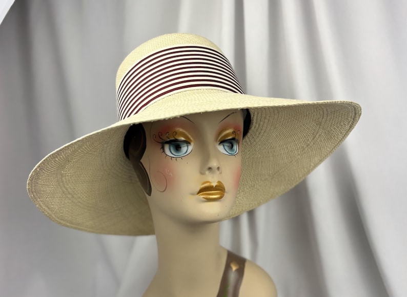 Pu&ograve; includere: Un cappello di paglia a tesa larga con corona e tesa color crema. Il cappello presenta una fascia a strisce bordeaux e bianche alternate intorno alla corona. Il cappello &egrave; esposto su una testa di manichino.