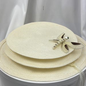 May include: A set of three stacked circular cream-colored woven placemats. The top placemat features a decorative dragonfly ornament with cream and brown wings and a small white bead head. The woven texture is consistent across all three pieces.