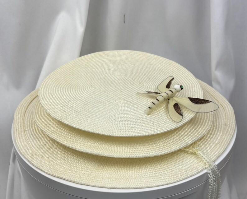 May include: A set of three stacked circular cream-colored woven placemats. The top placemat features a decorative dragonfly ornament with cream and brown wings and a small white bead head. The woven texture is consistent across all three pieces.