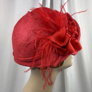 May include: A bright red hat constructed from a woven straw-like material, featuring a structured dome shape. The hat is adorned with a cluster of matching red satin fabric flowers and delicate, wispy feathers attached to the side of the crown.