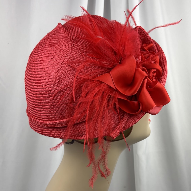 May include: A bright red hat constructed from a woven straw-like material, featuring a structured dome shape. The hat is adorned with a cluster of matching red satin fabric flowers and delicate, wispy feathers attached to the side of the crown.