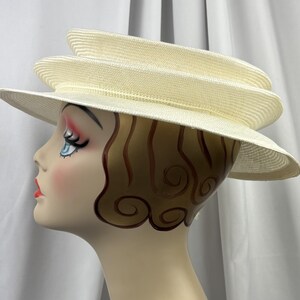 May include: A cream-colored straw hat featuring a multi-tiered, layered crown design with a wide, slightly downturned brim. The hat is displayed on a mannequin head with stylized brown hair, blue eyes, and red lips, showcasing the hat's structure.