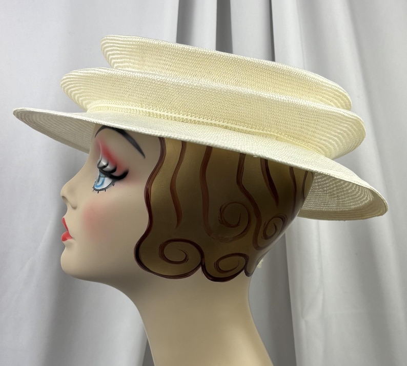 May include: A cream-colored straw hat featuring a multi-tiered, layered crown design with a wide, slightly downturned brim. The hat is displayed on a mannequin head with stylized brown hair, blue eyes, and red lips, showcasing the hat's structure.