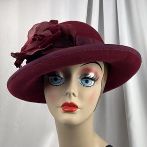 May include: A deep burgundy felt hat featuring a wide brim and a matching ribbon band. The hat is adorned with a large, layered fabric flower on the side, crafted from sheer and velvet-textured material in a coordinating dark red shade.
