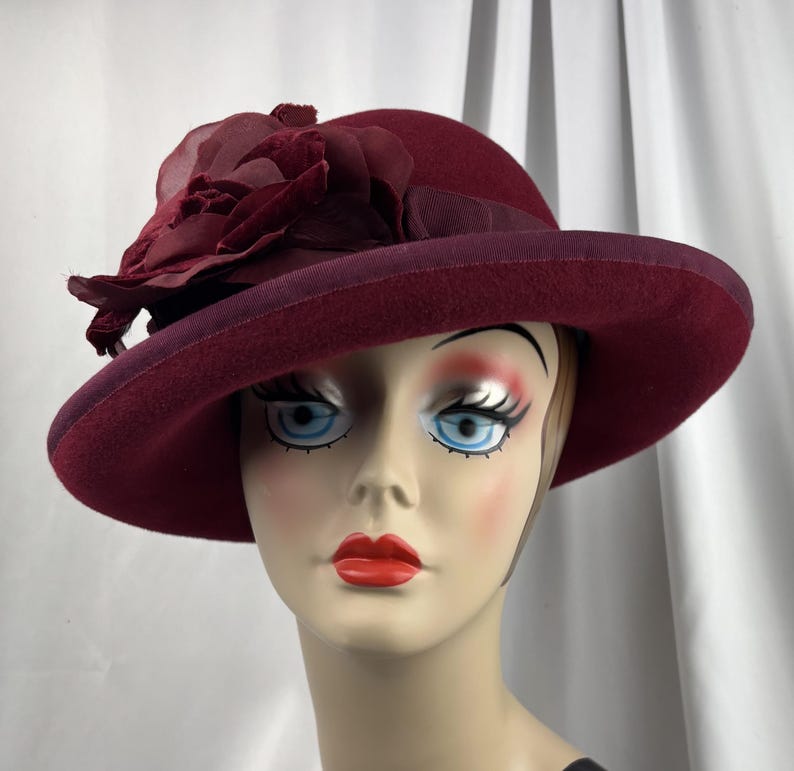 May include: A deep burgundy felt hat featuring a wide brim and a matching ribbon band. The hat is adorned with a large, layered fabric flower on the side, crafted from sheer and velvet-textured material in a coordinating dark red shade.
