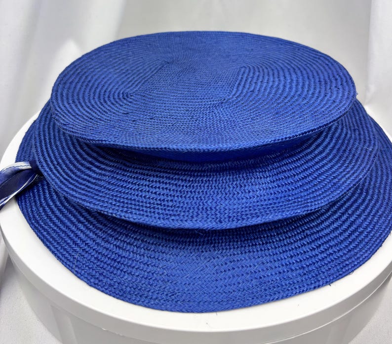 K&ouml;nnte beinhalten: Ein leuchtend k&ouml;nigsblauer Strohhut mit einer breiten, flachen Krempe und einer strukturierten, runden Krone. Der Hut besteht aus eng geflochtenem Strohmaterial und verf&uuml;gt &uuml;ber ein kleines, passendes blaues Band an der Seite der Krempe.