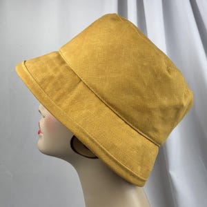 Puede incluir: Un sombrero de pescador color amarillo mostaza con una copa estructurada y un ala inclinada hacia abajo con detalles de costuras horizontales visibles. El sombrero est&aacute; confeccionado en tela de color s&oacute;lido con una textura sutil y dise&ntilde;o simple.