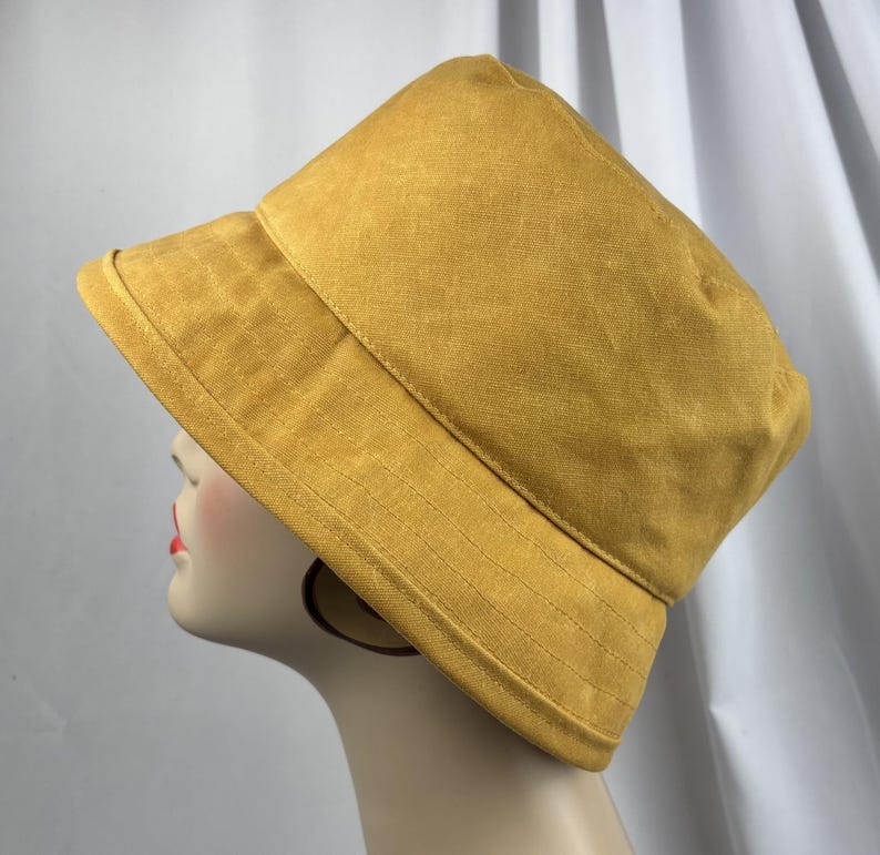 Puede incluir: Un sombrero de pescador color amarillo mostaza con una copa estructurada y un ala inclinada hacia abajo con detalles de costuras horizontales visibles. El sombrero est&aacute; confeccionado en tela de color s&oacute;lido con una textura sutil y dise&ntilde;o simple.