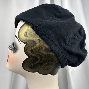 May include: A solid black beret featuring visible decorative stitching along the crown and the lower edge. The hat is displayed on a mannequin head with stylized gold-toned hair and long eyelashes, showcasing the rounded shape of the headwear.