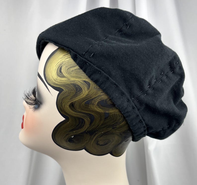 May include: A solid black beret featuring visible decorative stitching along the crown and the lower edge. The hat is displayed on a mannequin head with stylized gold-toned hair and long eyelashes, showcasing the rounded shape of the headwear.