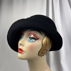 May include: A solid black felt hat with a wide, slightly downturned brim and a rounded crown. The hat is displayed on a mannequin head featuring painted facial details, including blue eyes, red lips, and stylized gold hair accents.