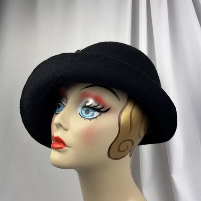 May include: A solid black felt hat with a wide, slightly downturned brim and a rounded crown. The hat is displayed on a mannequin head featuring painted facial details, including blue eyes, red lips, and stylized gold hair accents.