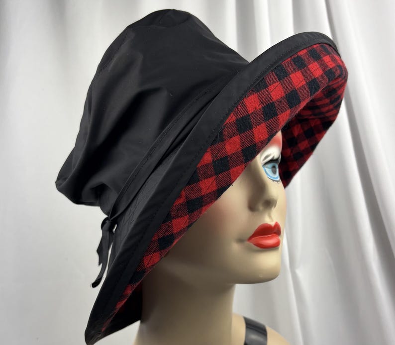 Pu&ograve; includere: Cappello da sole nero con tesa larga. Il lato inferiore della tesa presenta un motivo a quadri rossi e neri. Un nastro nero &egrave; legato sul lato del cappello. Il cappello &egrave; esposto su una testa di manichino.