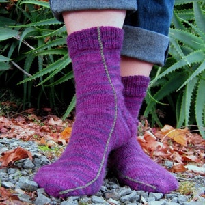 May include: A pair of purple and green striped socks with a contrasting green seam down the back of each sock. The socks are worn by a person standing on a bed of gravel and leaves.