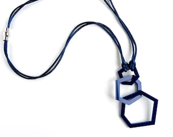 Può includere: Una collana blu navy con tre ciondoli esagonali intrecciati di dimensioni variabili, sospesi a un cordino a doppio filo. Il cordino include una chiusura magnetica cilindrica color argento e i ciondoli geometrici sono realizzati in materiale liscio.