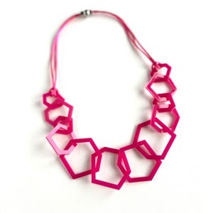 May include: A necklace featuring a series of interconnected hot pink hexagonal frames of varying sizes. The geometric shapes are linked together to form a cascading pattern, suspended from a thin, matching pink cord with a small metallic clasp.